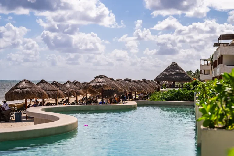 A beach side swim up pool with palapas and ocean view in the background.
